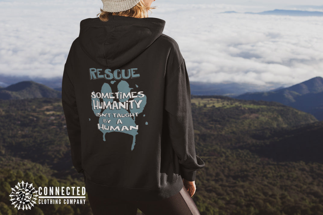 Model wearing Black Show Humanity Unisex Hoodie at the mountains that reads "Rescue. Sometimes humanity isn't taught by a human" - marktwainstoryteller - Ethically and Sustainably Made - 10% donated to the Society for the Prevention of Cruelty to Animals